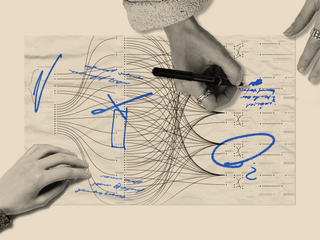 An overhead shot of hands writing on paper with blue marks annotating a drawing.